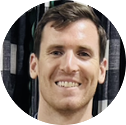 Smiling man with short brown hair and light skin in front of dark curtains.