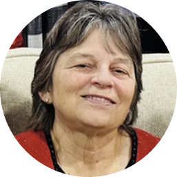 Smiling older woman with short gray hair wearing a red top and sitting on a beige couch.