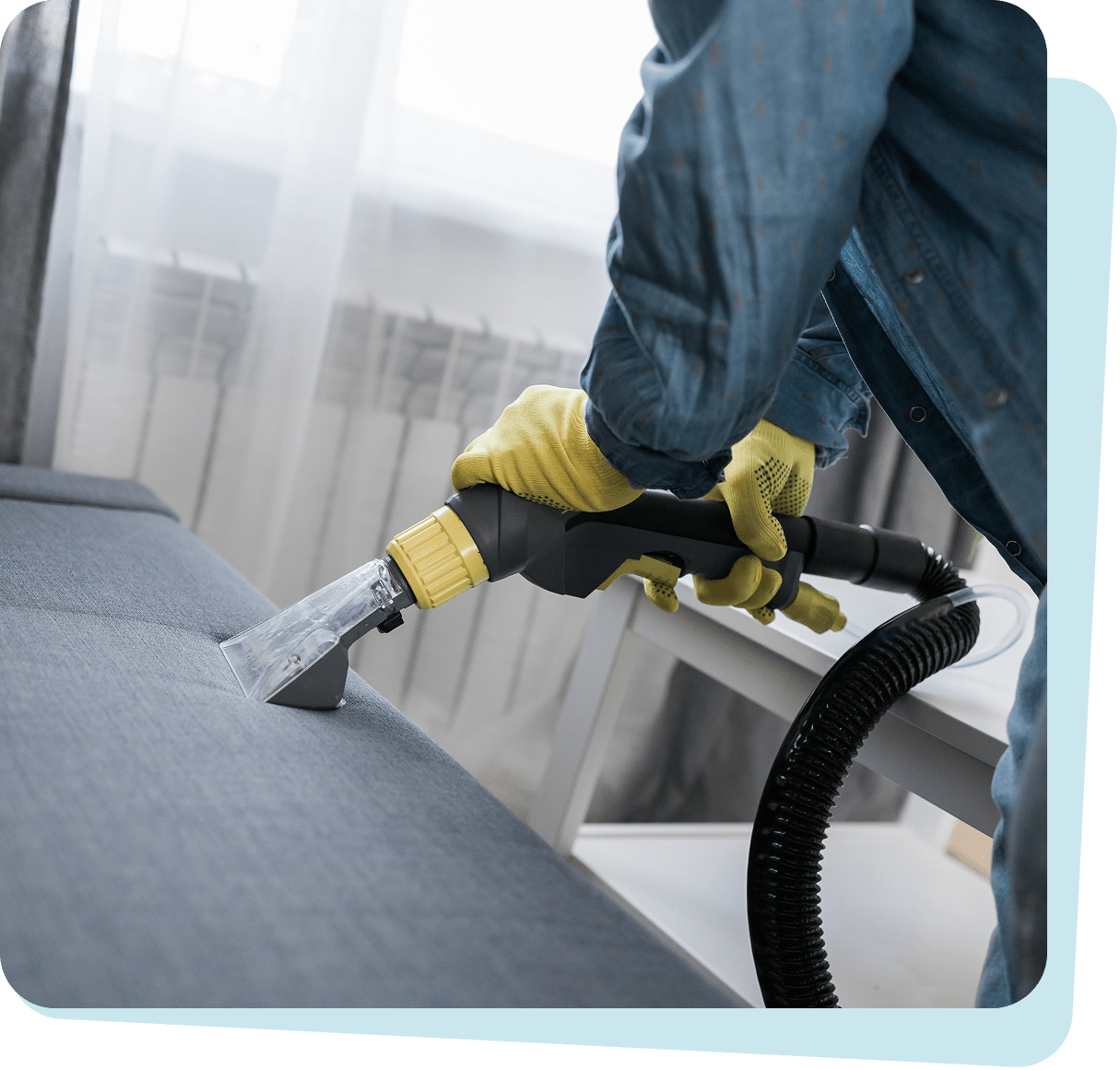 Person wearing yellow gloves using a vacuum upholstery cleaner on a gray sofa.