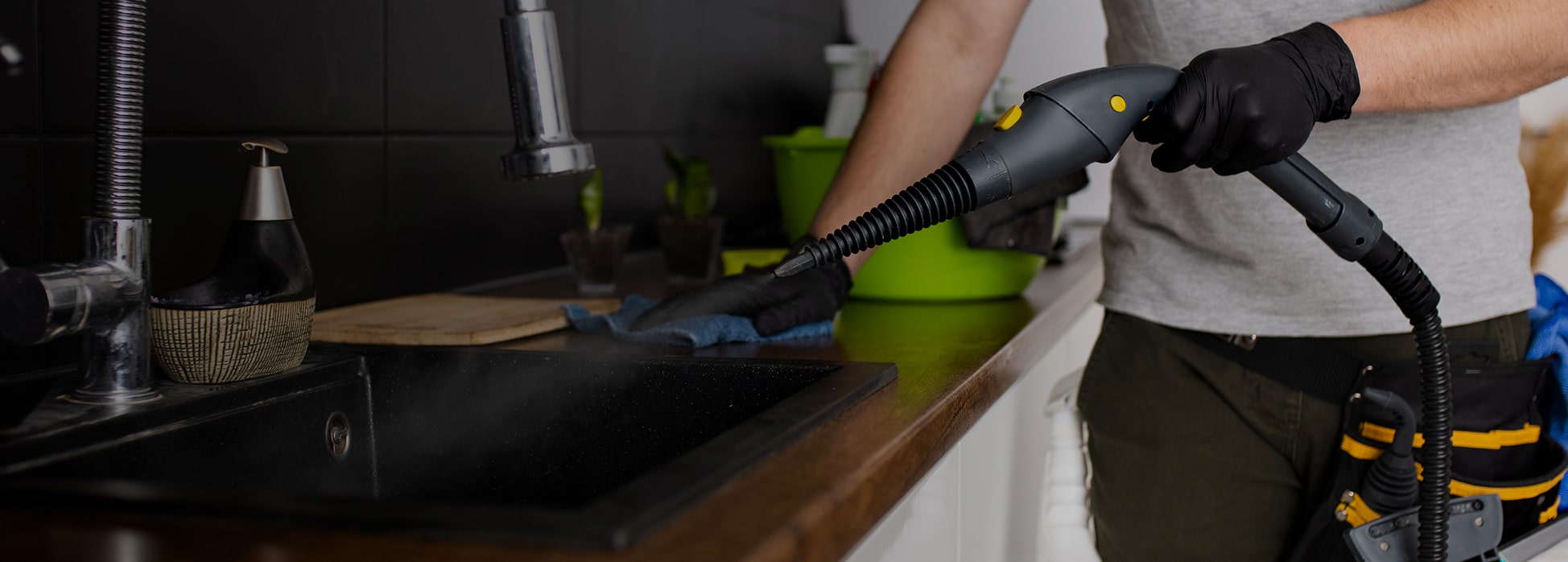 Person wearing black gloves cleaning a kitchen sink area with a steam cleaning device and a cloth.