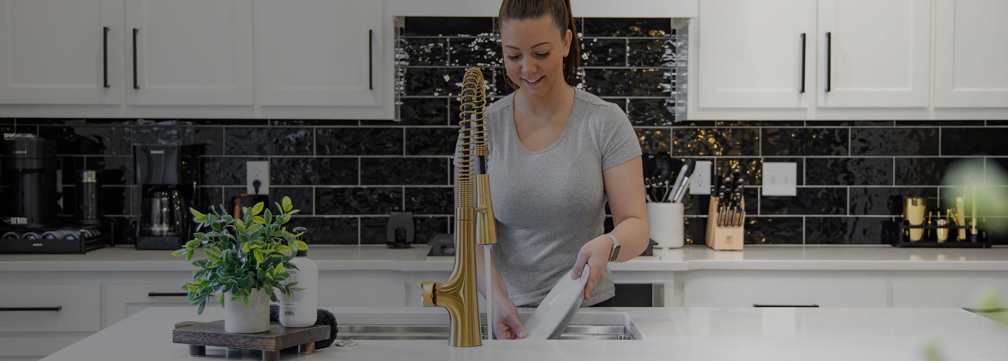 Woman washing a white plate at a kitchen sink with a gold faucet in a modern kitchen.