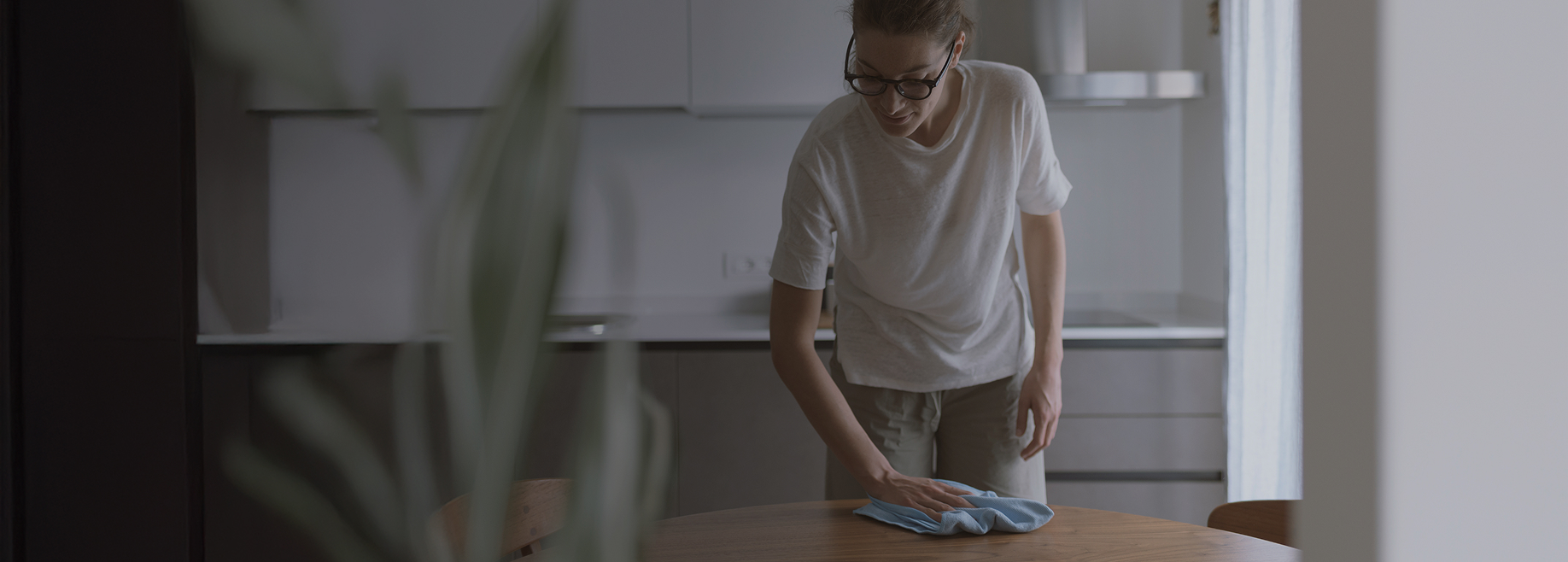 Woman wearing glasses cleaning a wooden kitchen table with a blue cloth.