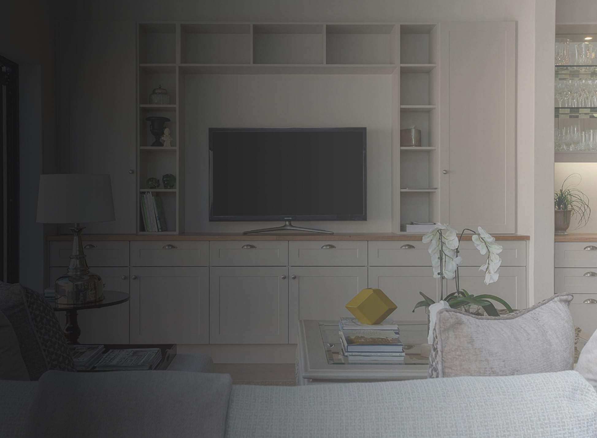 Modern living room with built-in white cabinetry surrounding a flat-screen TV, a glass coffee table with books and a yellow decorative object, and a gray sofa with a white orchid plant.