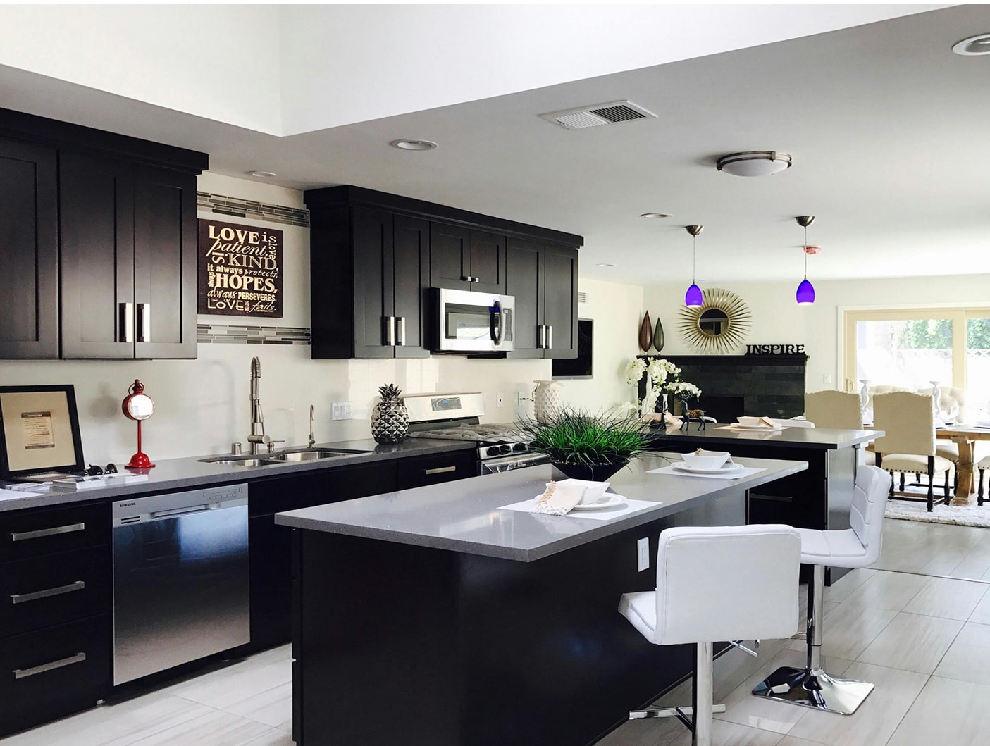 Modern kitchen with dark cabinets, gray countertops, stainless steel appliances, white bar stools, and a decorative sign reading 'LOVE is patient, LOVE is kind' on the wall.