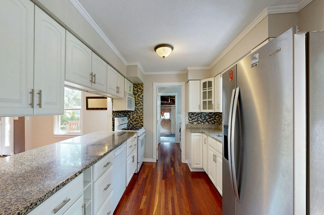 Narrow kitchen with white cabinets, granite countertops, stainless steel refrigerator, and wooden flooring.