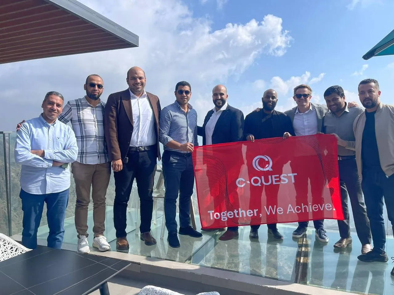 Group of nine men standing outdoors on a glass floor balcony under a partly cloudy sky, holding a red banner that reads 'C-QUEST Together, We Achieve.'