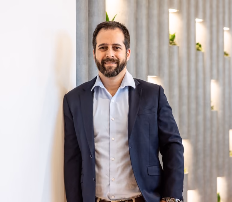 Kristopher Rezagholi, co-founder of legal tech software, ProPlaintiff.ai. Depicted with short dark hair and beard wearing a navy blazer and light blue shirt, standing against a wall with small illuminated plant niches.