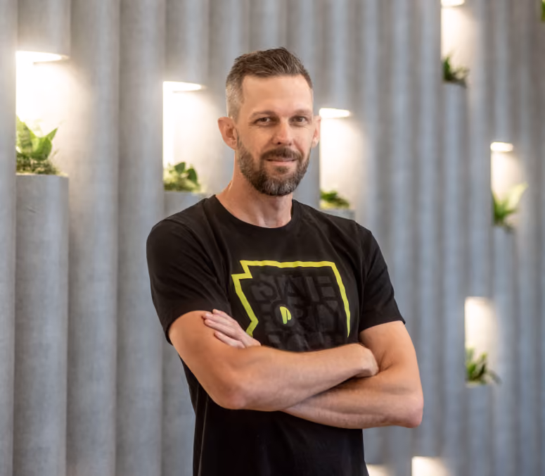 Jason Turnquist, co-founder of legal tech software, ProPlaintiff.ai. Depicted with short hair and beard wearing a black t-shirt, standing with arms crossed in front of a wall with small plants in lit niches.