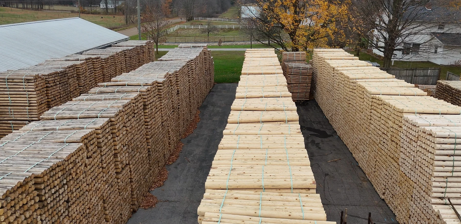 Large stacks of bundled wooden logs arranged in rows outdoors near a building and trees with autumn foliage.