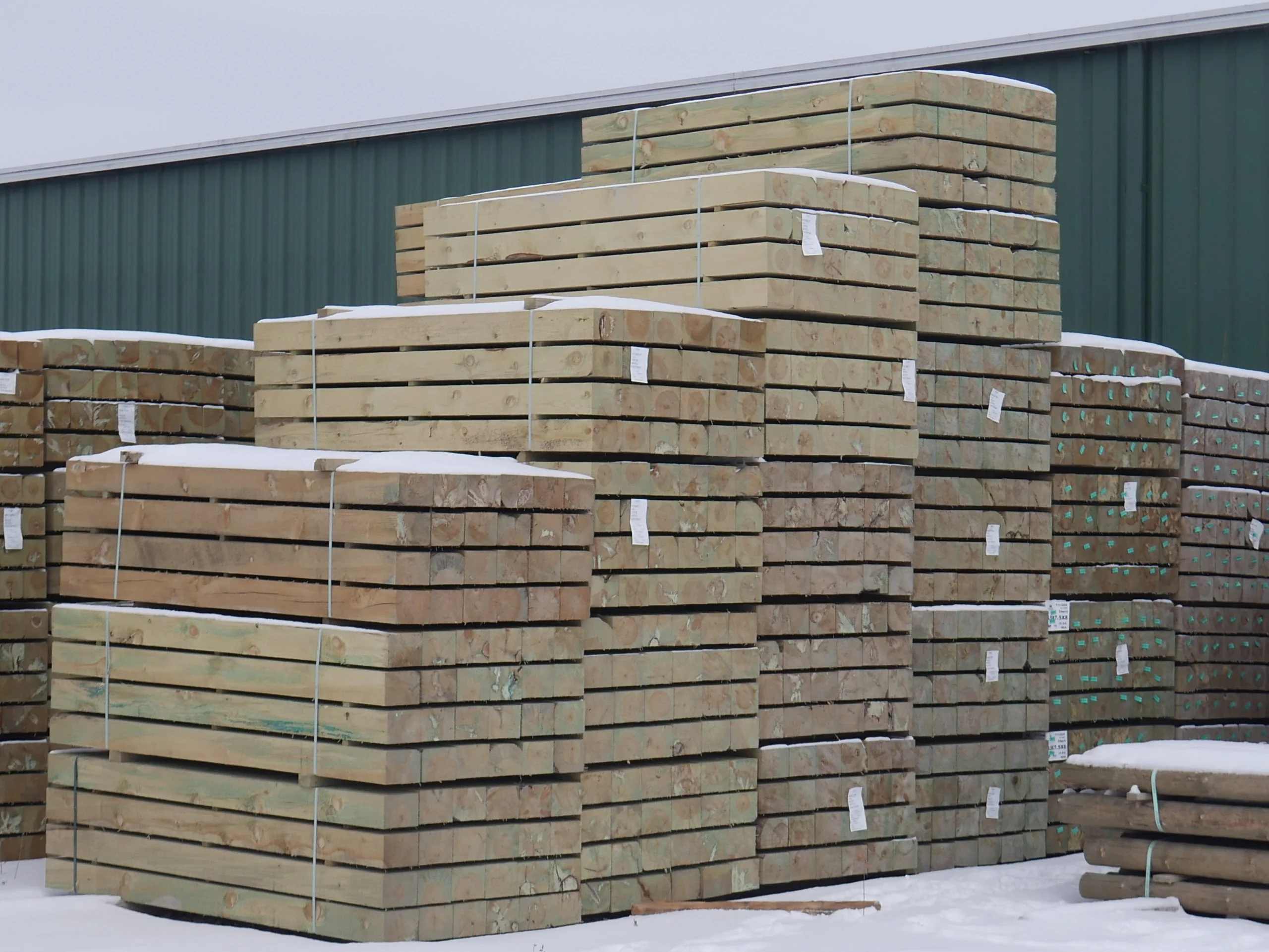 Stacks of bundled wooden planks and beams covered with snow outside a green building.