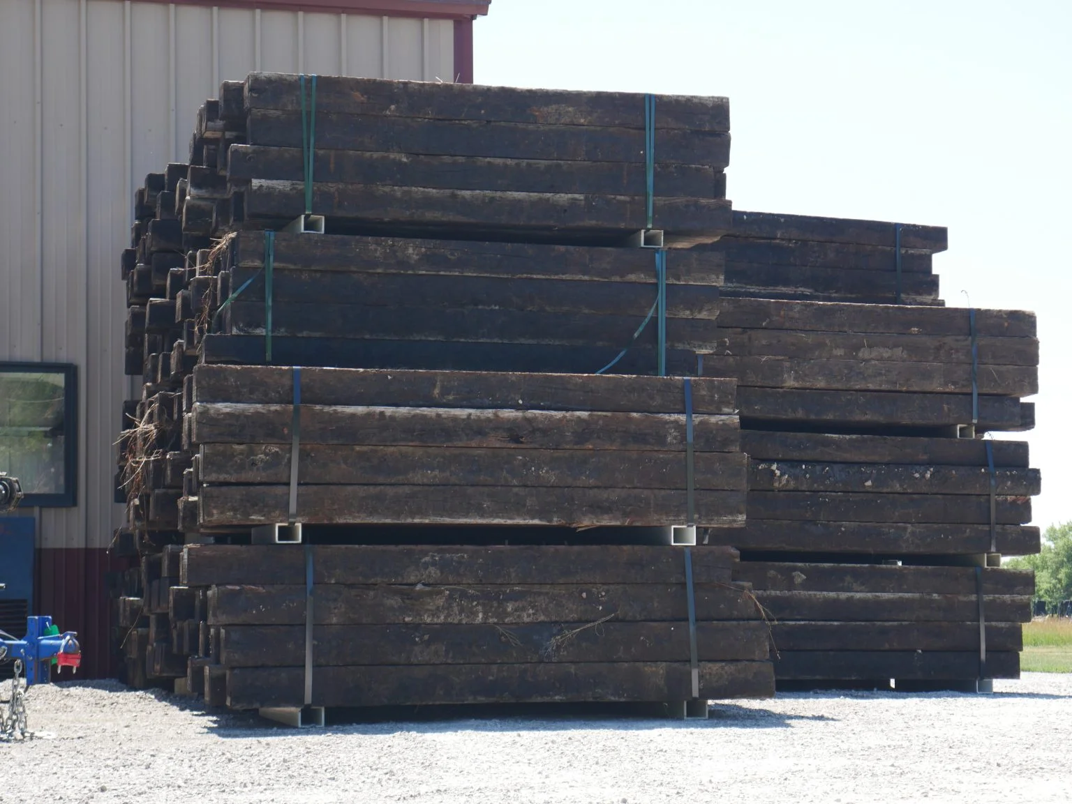 Stacks of dark, weathered wooden railroad ties bundled with metal straps outdoors on gravel near a building.