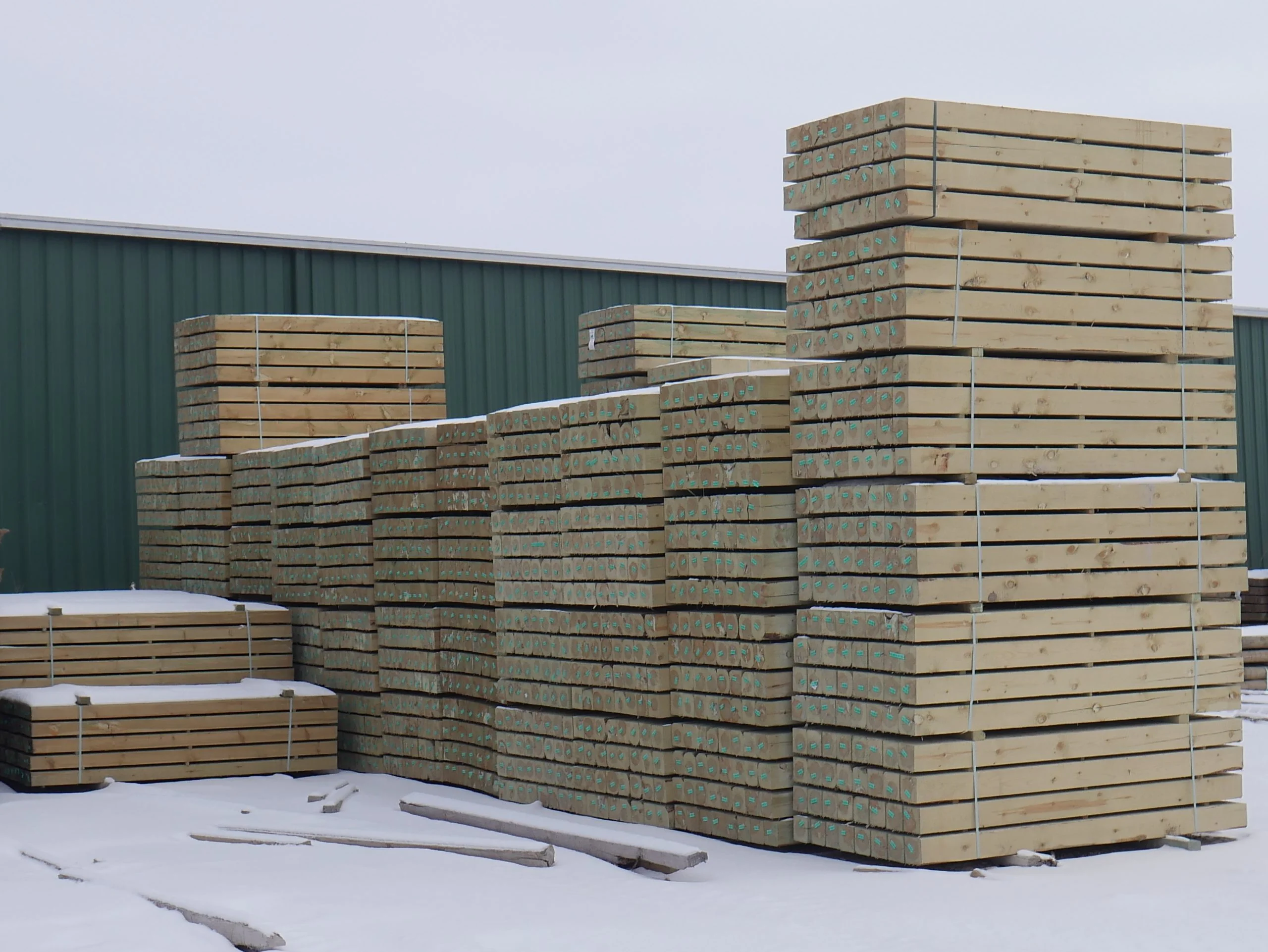 Stacks of wooden beams bundled and placed outdoors on snow-covered ground near a green metal building.