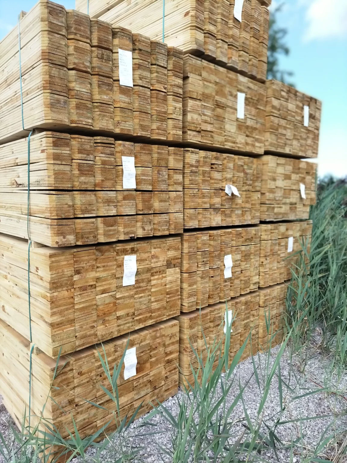 Stacks of neatly bundled, cut wooden planks outdoors on gravel with some tall grass nearby under a partly cloudy sky.