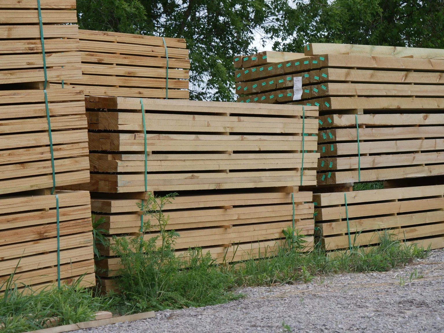 Stacks of cut lumber bundled and stored outdoors on gravel with green trees in the background.