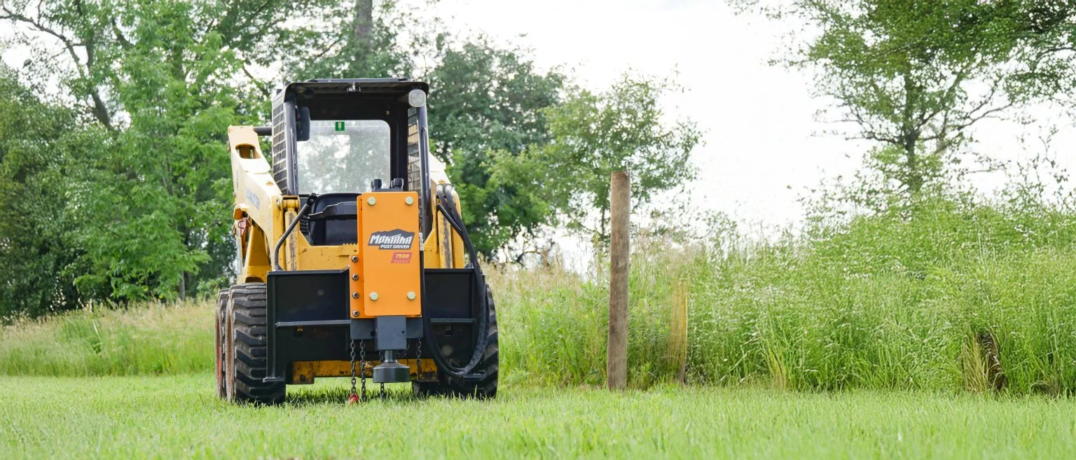 Yellow skid steer loader with a post driver attachment next to a wooden post on a grassy field.