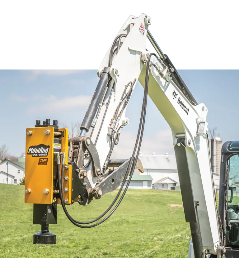 Close-up of a white Bobcat excavator arm equipped with a yellow Montana Post Driver attachment in a green field with farm buildings in the background.