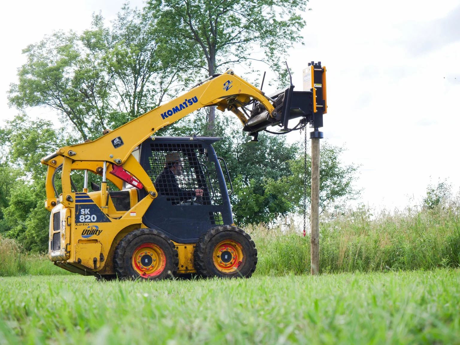 Yellow Komatsu skid-steer loader with post driver attachment installing a wooden post in grassy field.