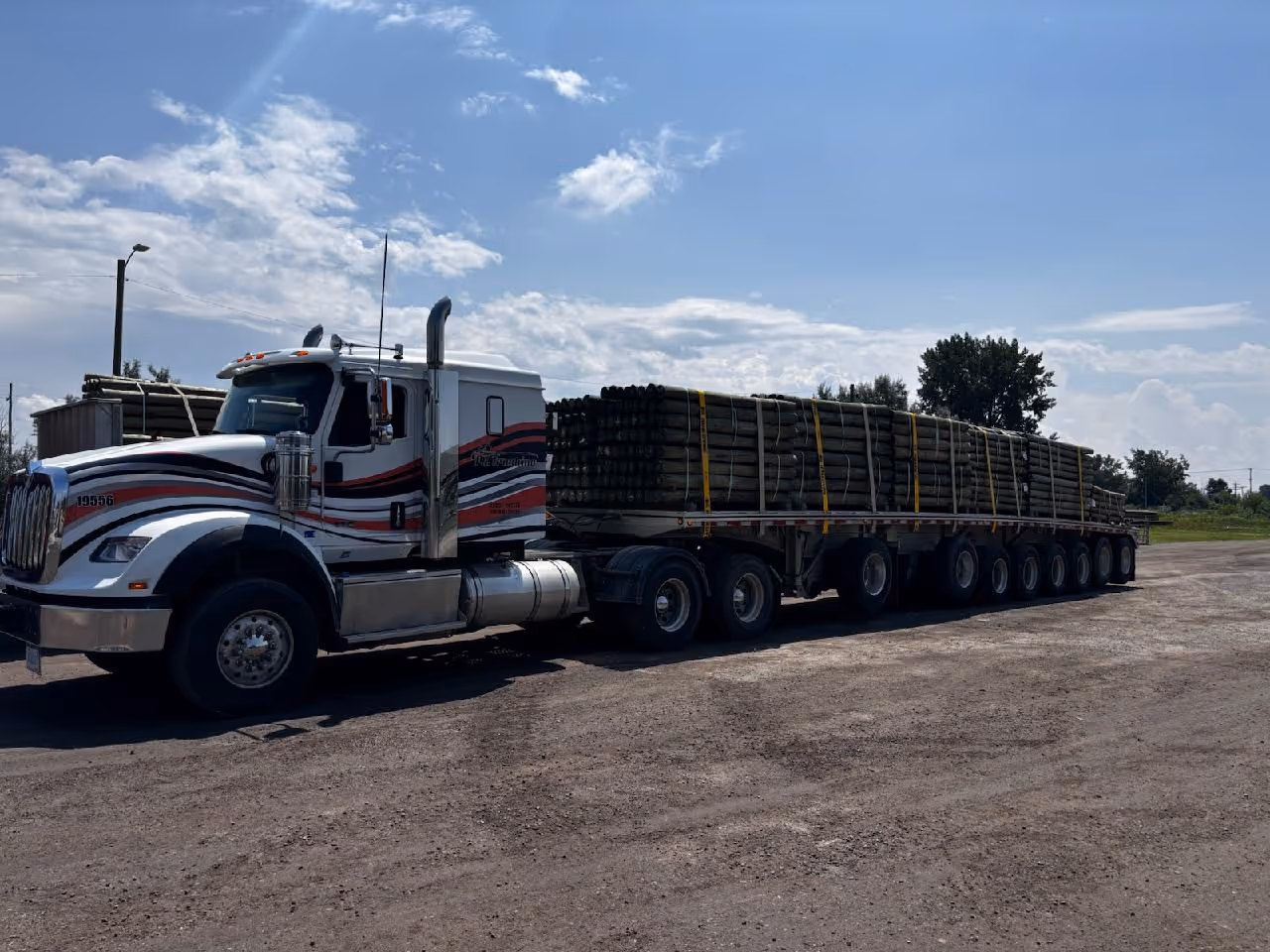 White semi-truck with red and black stripes hauling a long flatbed trailer loaded with secured pipes, parked on a gravel lot under a partly cloudy blue sky.