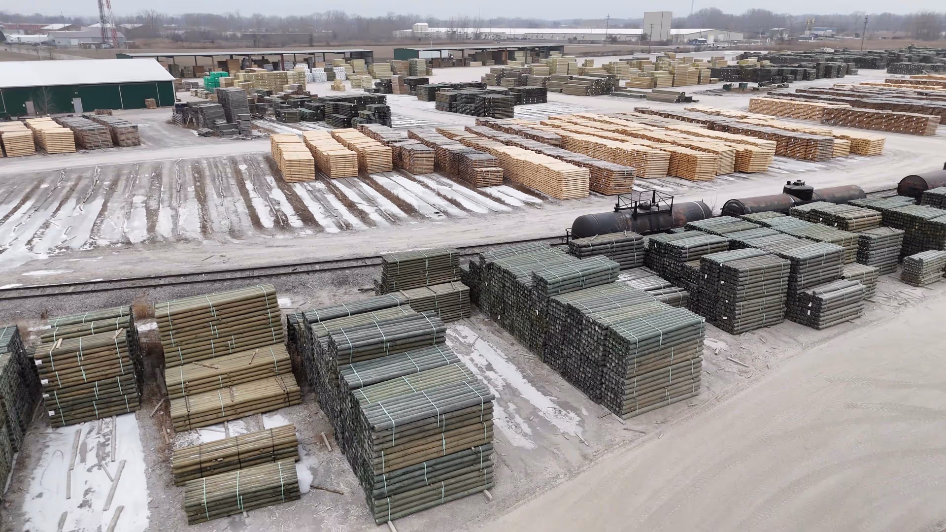 Large outdoor lumber yard with stacks of treated wooden poles and planks organized in rows on a snowy ground.