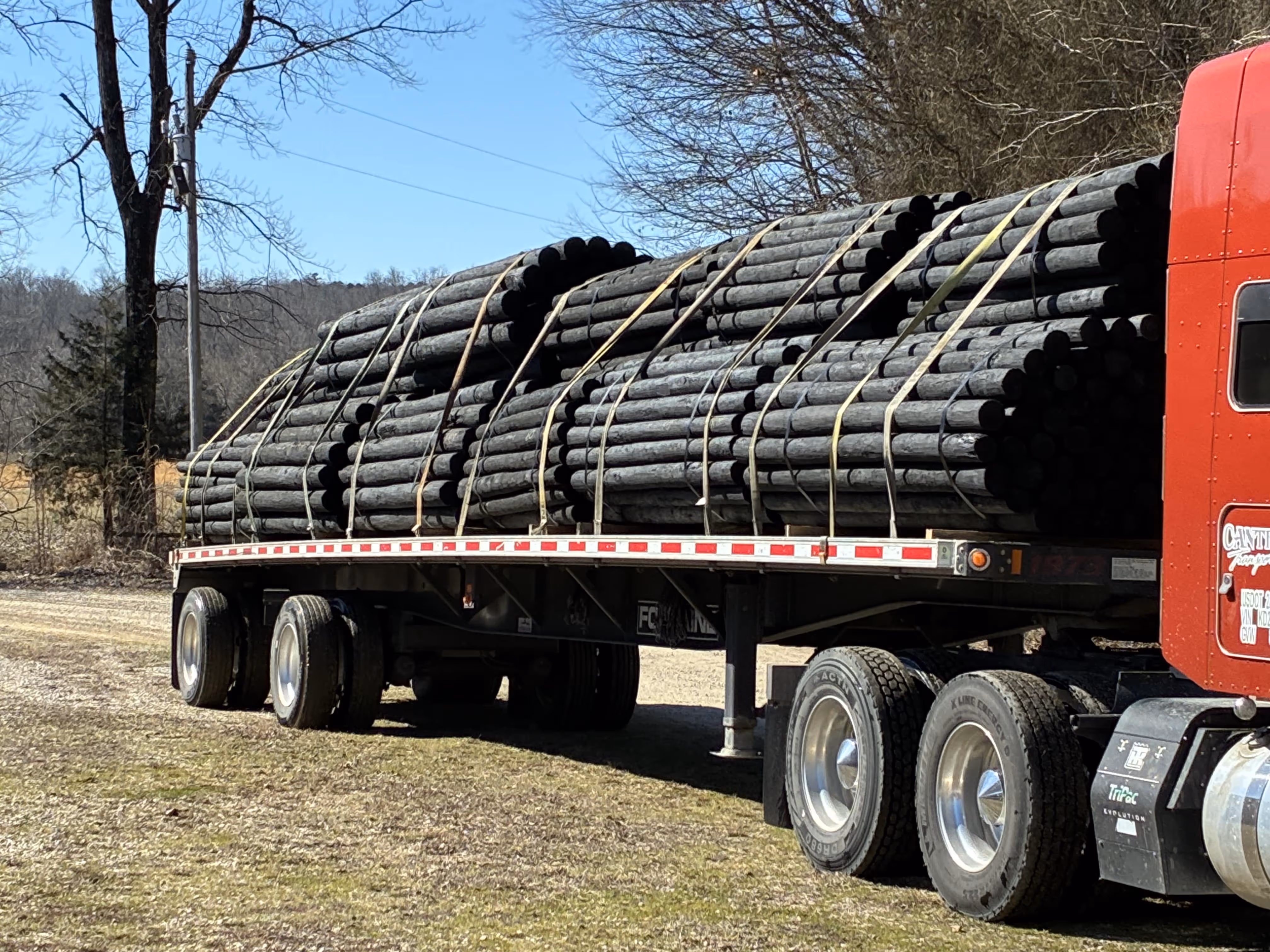 Flatbed truck carrying large bundles of dark creosote-coated wooden posts secured with straps.