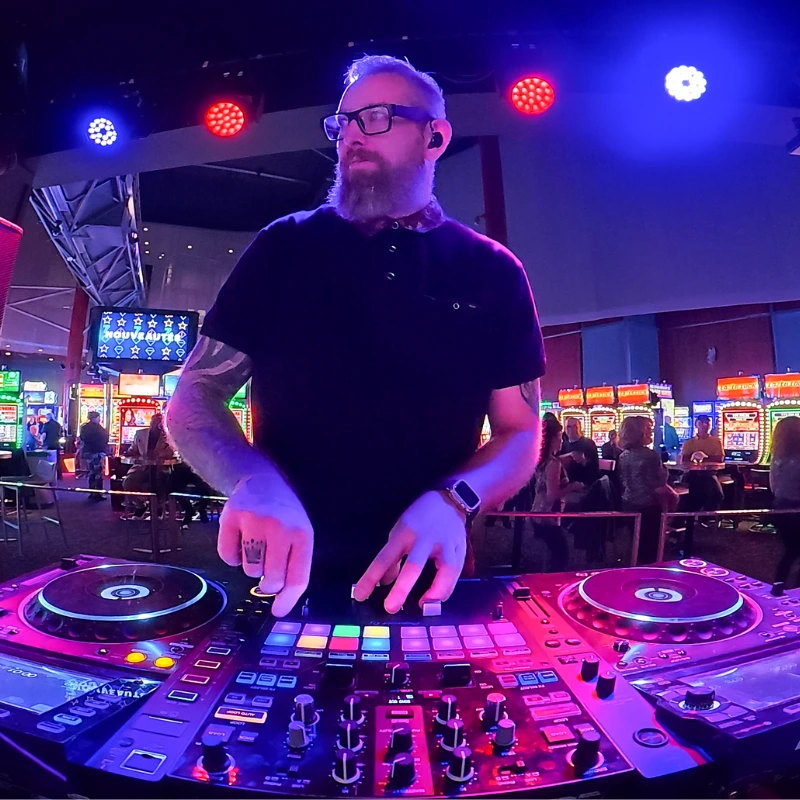 A bearded DJ wearing glasses and a black shirt stands behind a mixer with colorful lights, performing at lively Ottawa weddings in a casino filled with slot machines and guests enjoying premium DJ services.