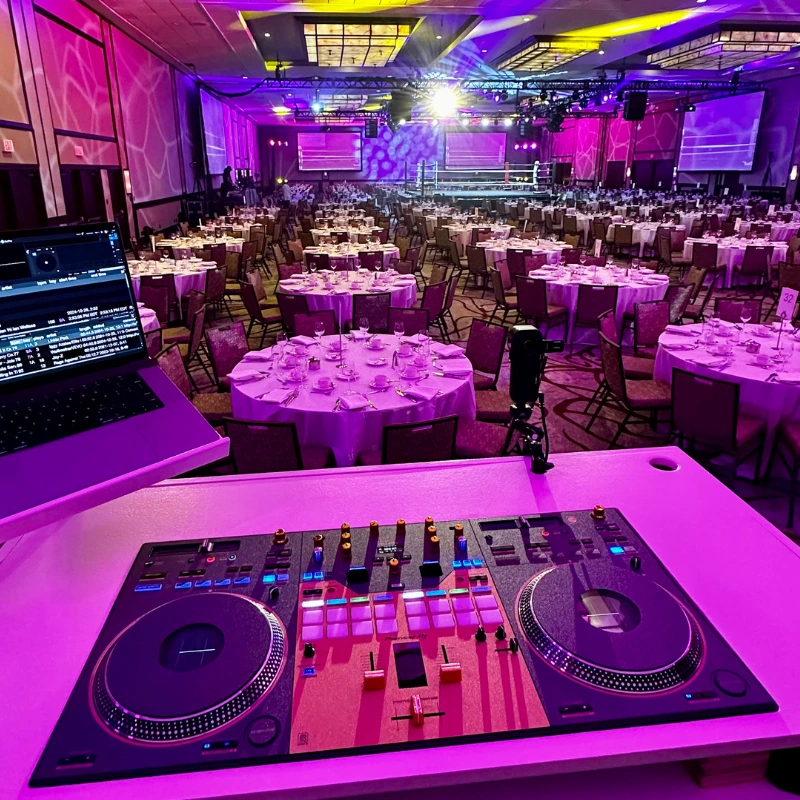 A DJ booth with a mixer and laptop overlooks a large, elegant banquet hall in Ottawa set for weddings or events, with round tables, chairs, and colorful stage lights illuminating the room—perfect for professional DJ services.