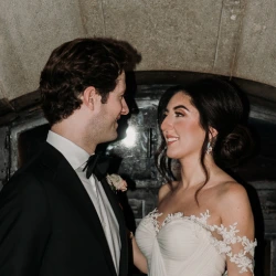 A bride in an off-shoulder white gown holding a colorful bouquet smiles at her groom in a black suit. They stand close together in front of a dark, arched background, sharing a loving gaze at their Ottawa wedding.