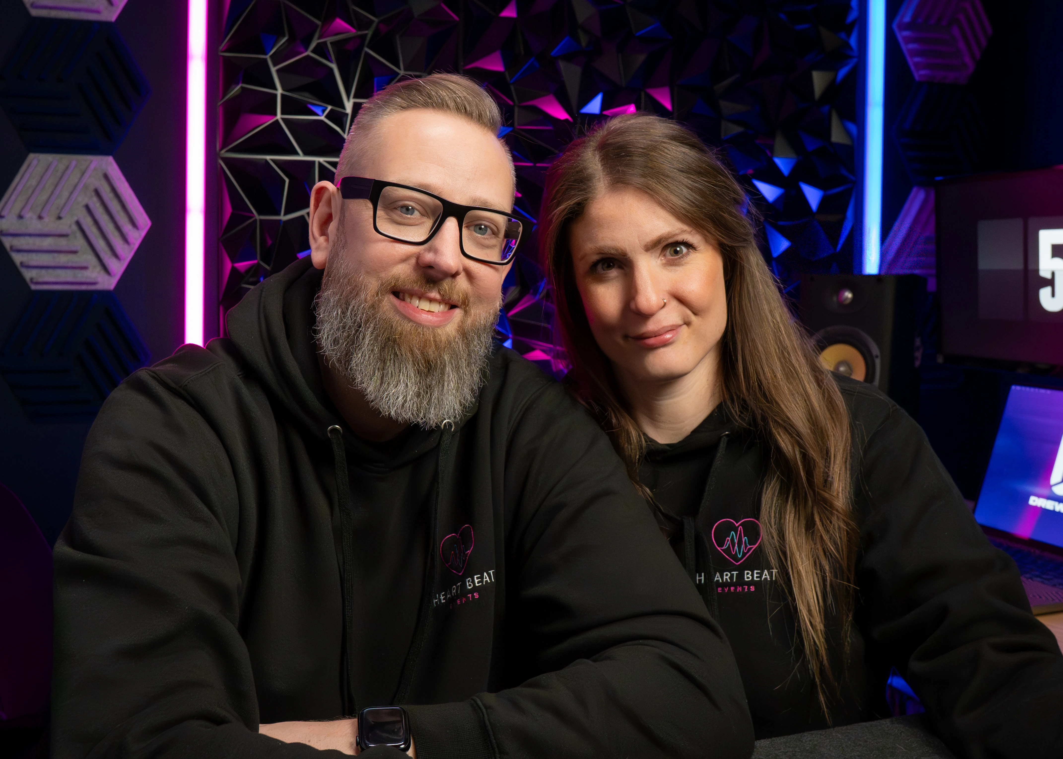 Smiling man with glasses and beard and woman with long hair wearing matching black hoodies in a colorful studio setting.