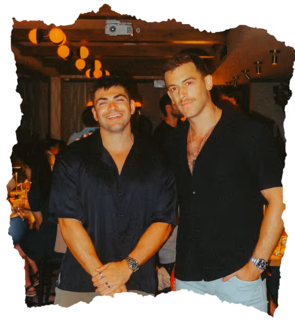 Two men wearing black shirts standing close together in a warmly lit indoor setting, smiling at the camera.