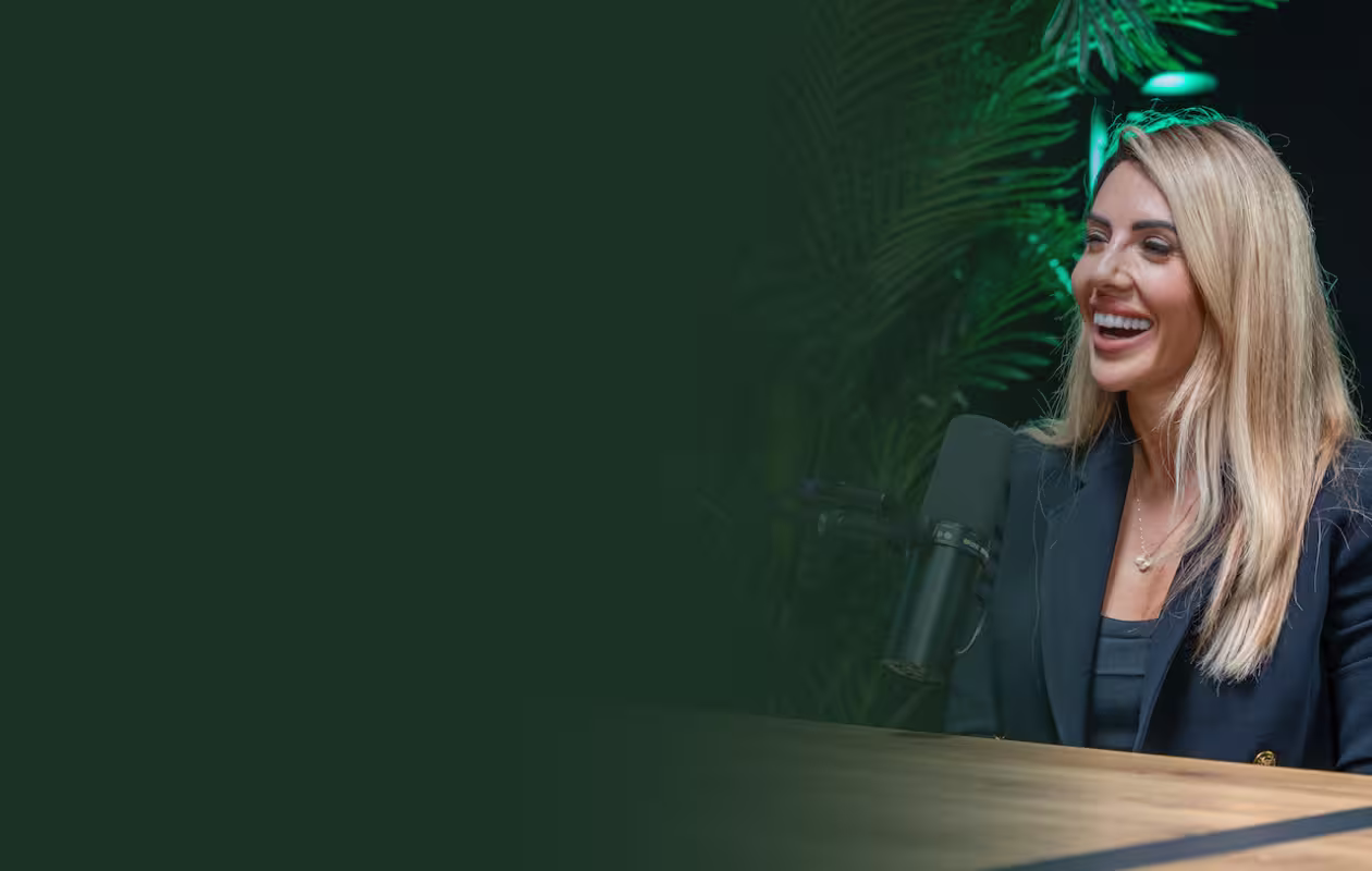 Smiling blonde woman speaking into a microphone while seated at a wooden table with green plants in the background.