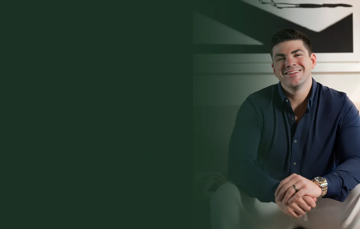 Smiling blonde woman speaking into a microphone while seated at a wooden table with green plants in the background.