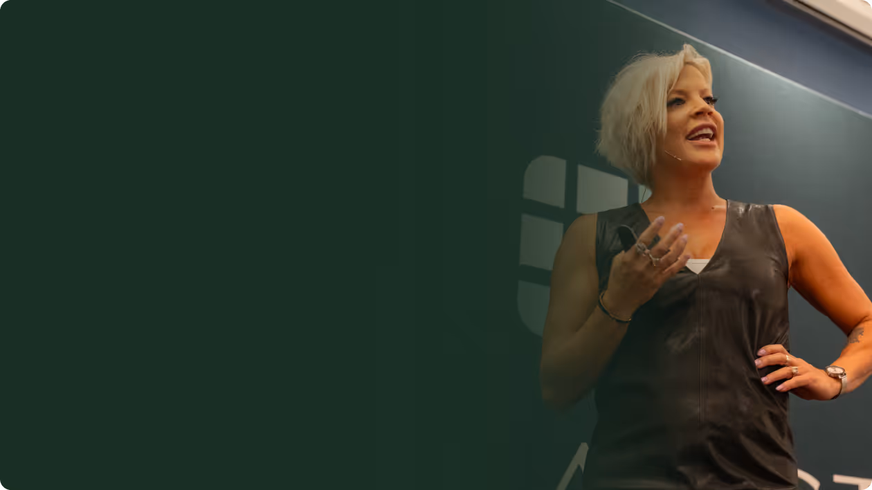 Man with white beard and black cap speaking into a microphone against a brown wall.