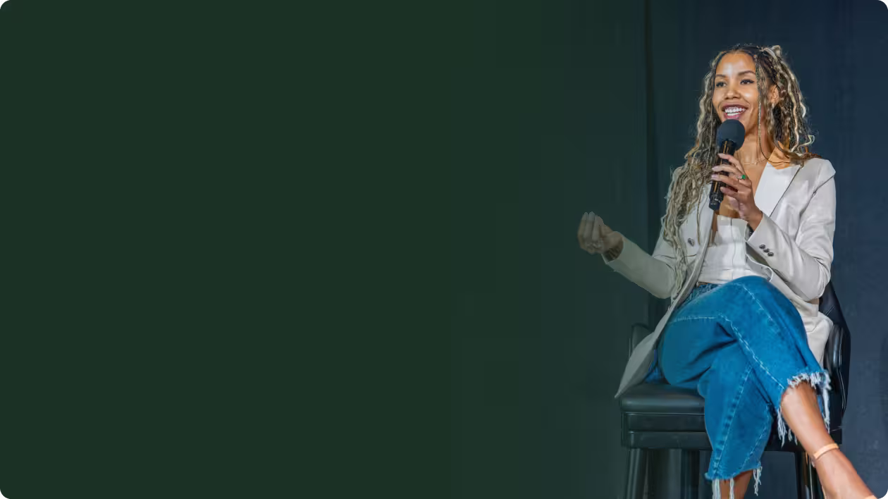 Man with white beard and black cap speaking into a microphone against a brown wall.