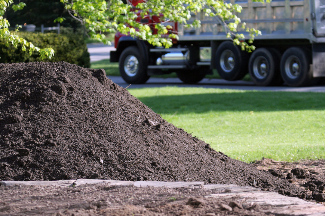 Pile of dark soil in a garden with a red dump truck and green grass in the background.