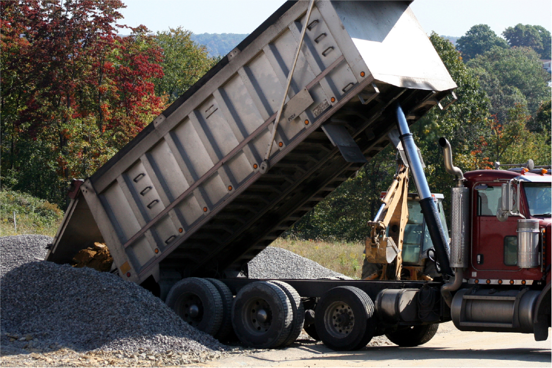 Dump truck unloading gravel or rocks at a construction site with trees in the background.