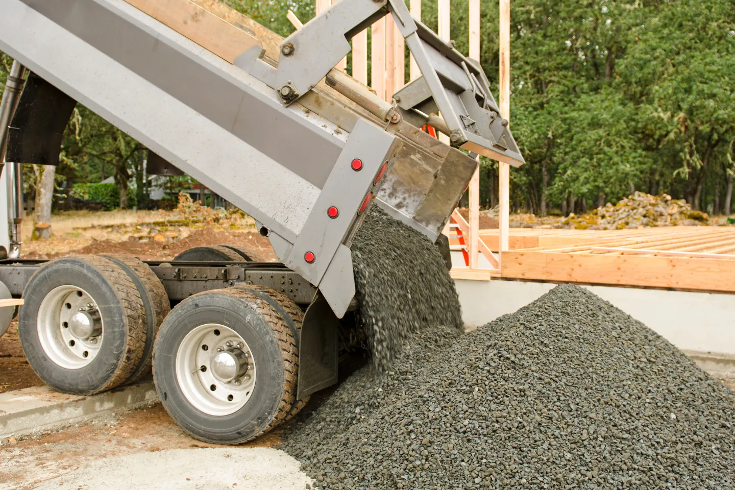 Dump truck unloading gravel at a construction site with wooden framing and trees in the background.