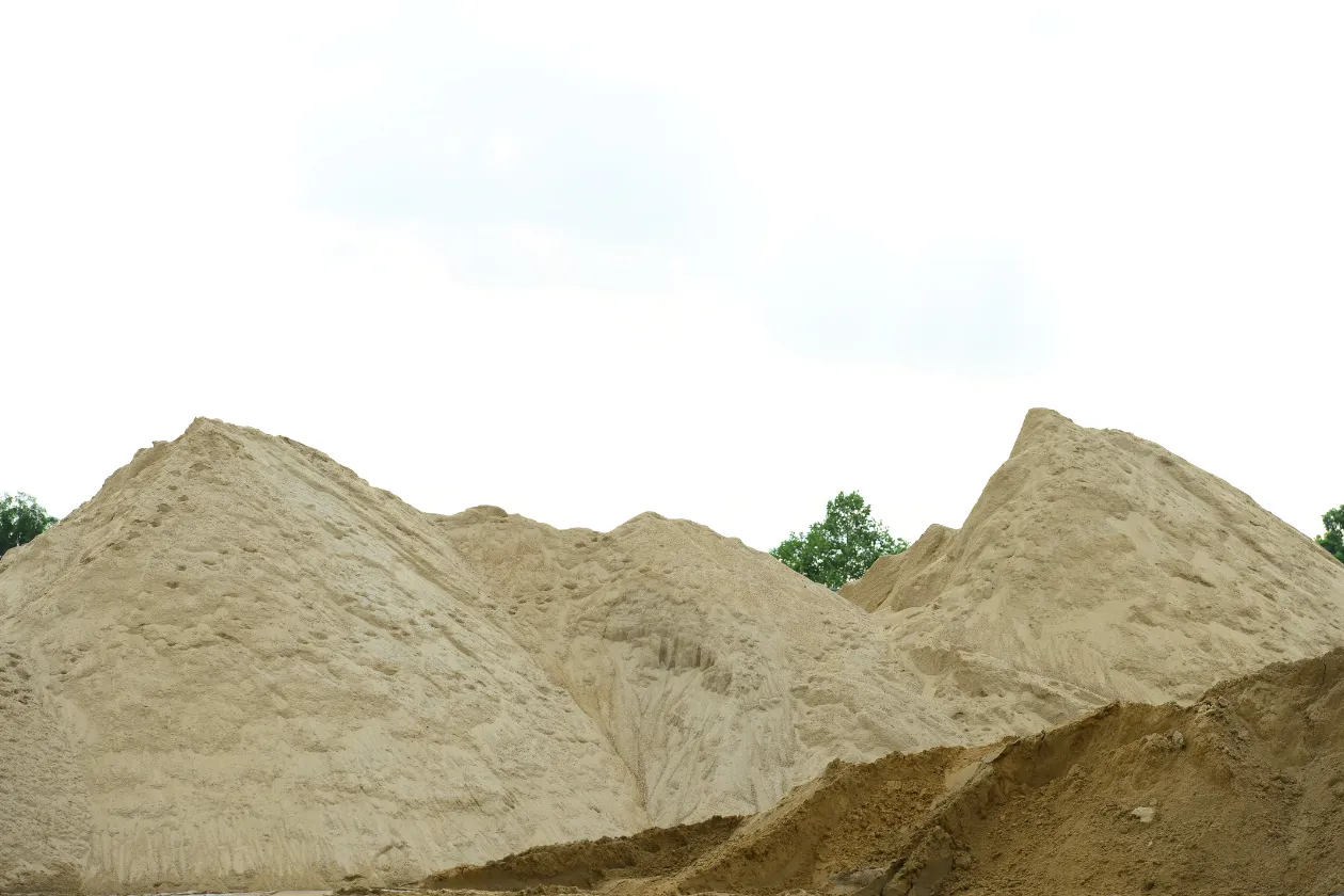 Several large piles of sand with trees visible in the background under a bright, overcast sky.