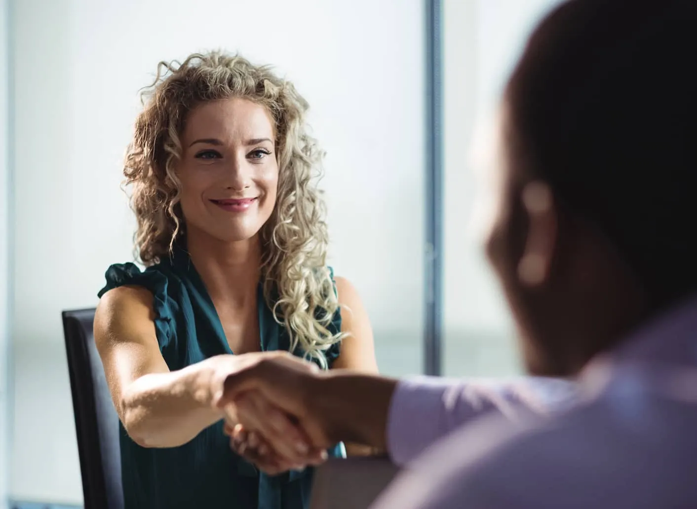 Smiling woman with curly hair shaking hands with a person in a purple shirt during a meeting.