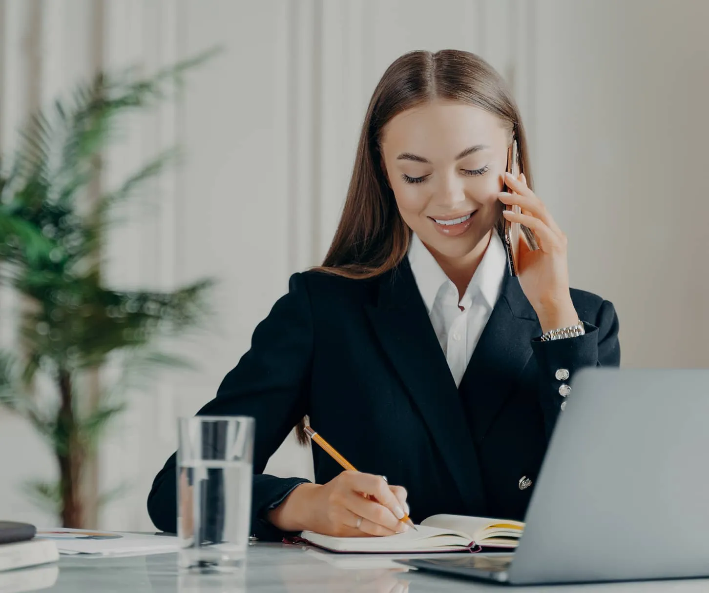 Smiling businesswoman in a black blazer talking on a smartphone while writing in a notebook at a desk with a laptop and a glass of water.