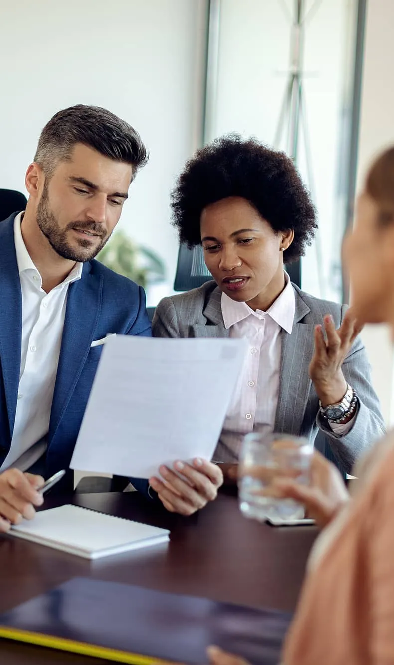 Two business professionals reviewing a document while a third person holds a glass of water nearby.