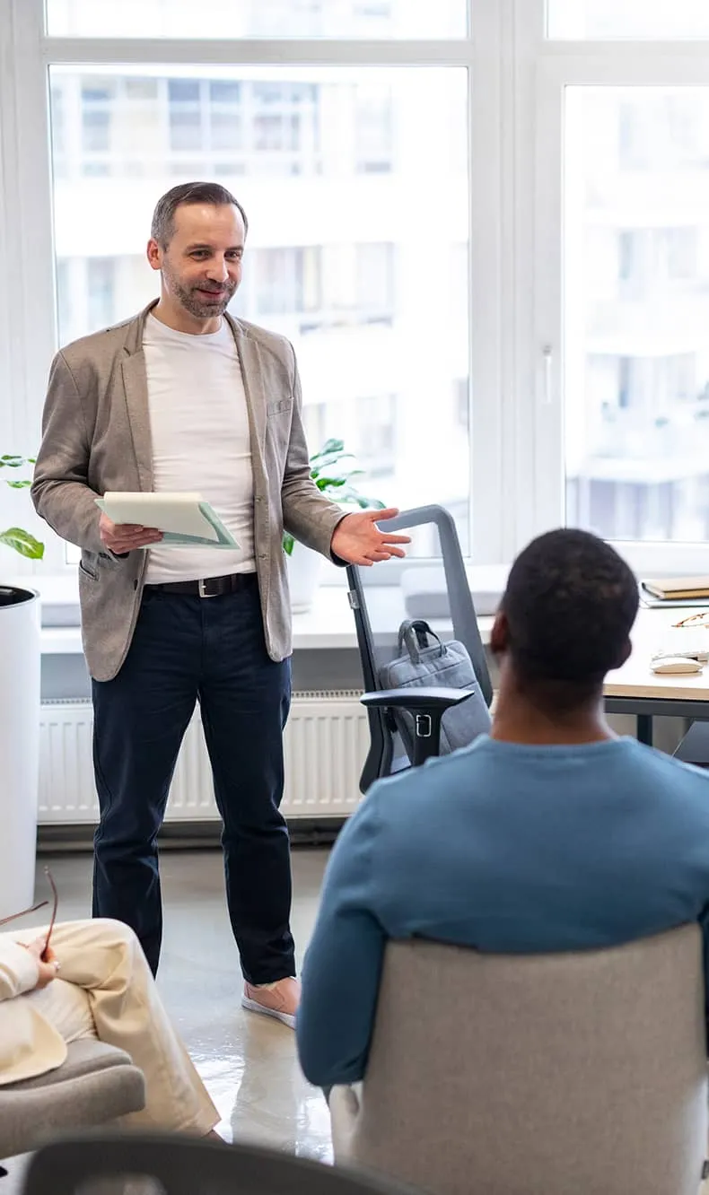 Man in a blazer holding a clipboard speaking to seated colleagues in a bright modern office.