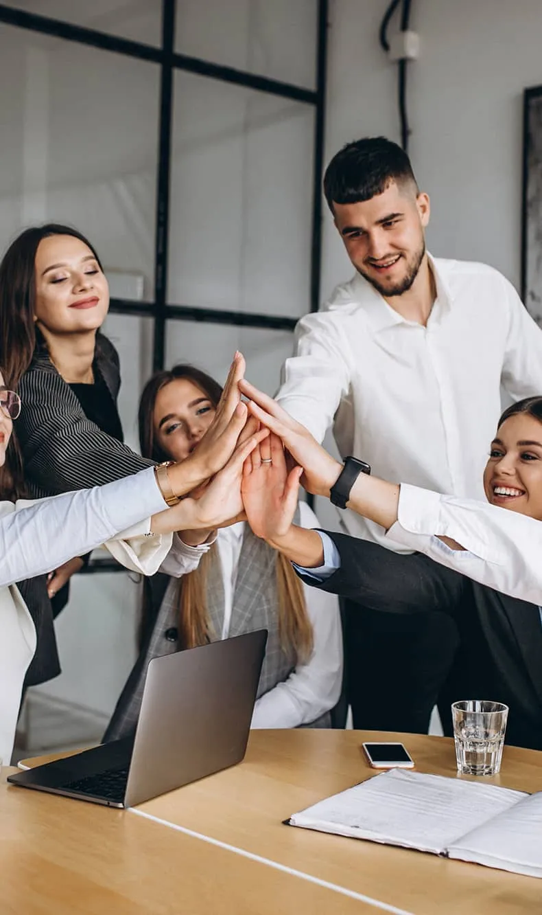 A group of young professionals in business attire giving a collective high-five around a conference table with a laptop, notebook, phone, and glass of water.