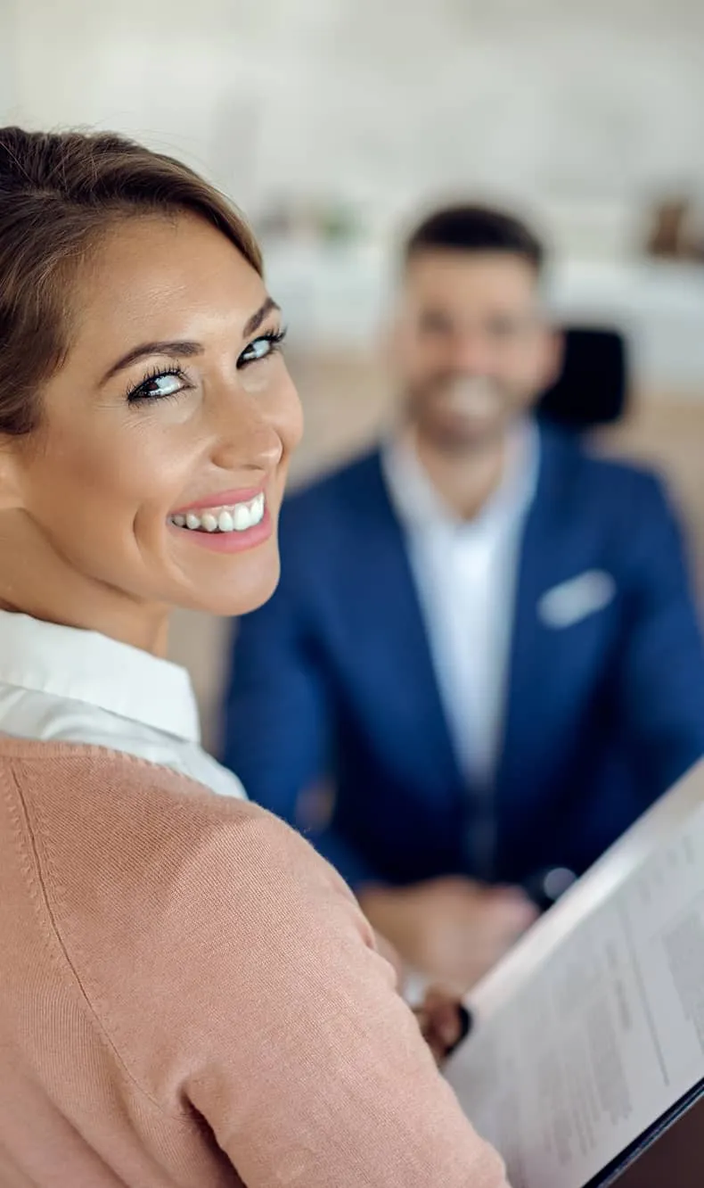 Smiling woman holding a clipboard with a man in a blue suit blurred in the background.