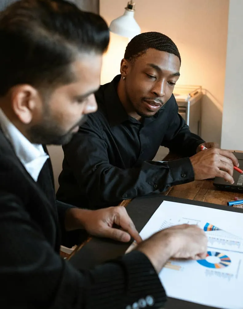 Two men collaborating over charts and graphs on a desk, one pointing and the other holding a pen near a laptop.
