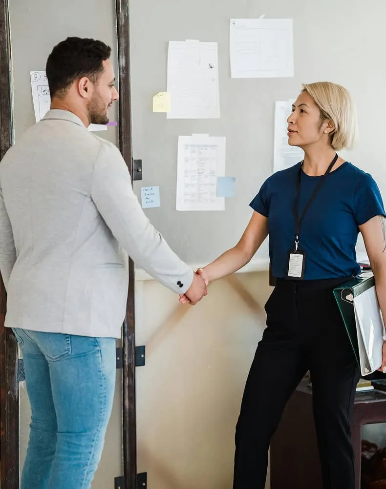 Man in light blazer and jeans shaking hands with woman in navy shirt and black pants holding a folder in an office room with papers on the wall.