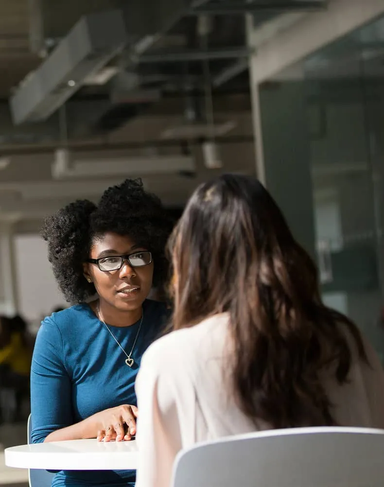 Two women having a focused conversation across a white table in a modern office setting.