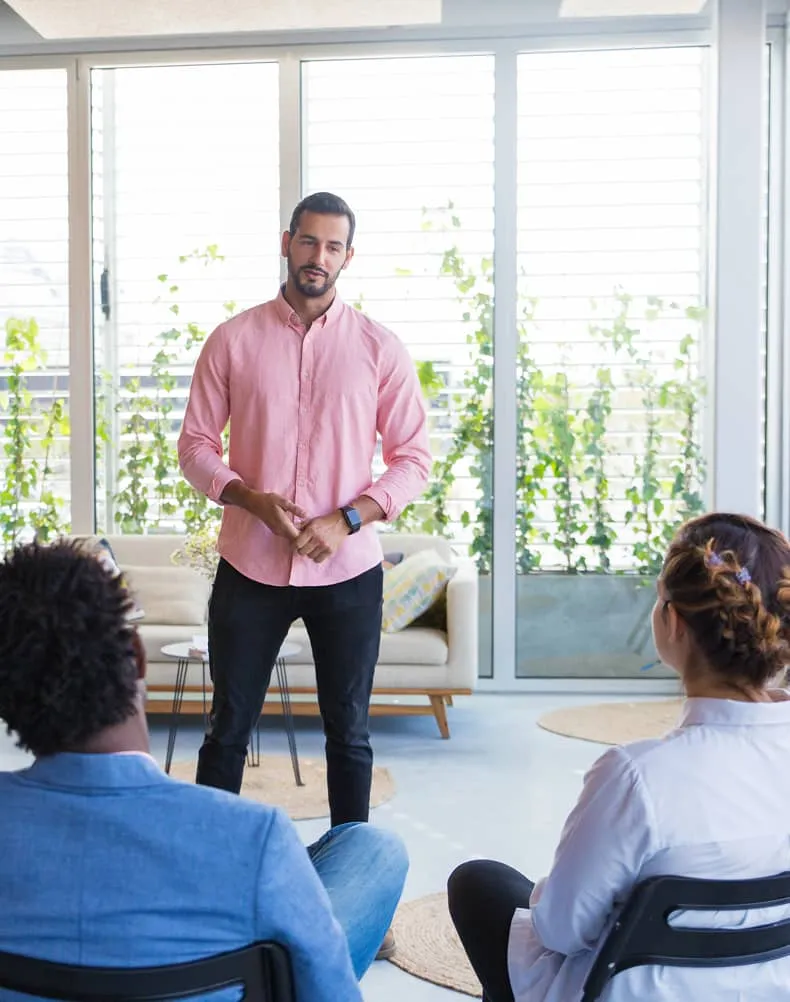 Man in pink shirt standing and speaking to two seated listeners in a bright room with large windows and plants outside.