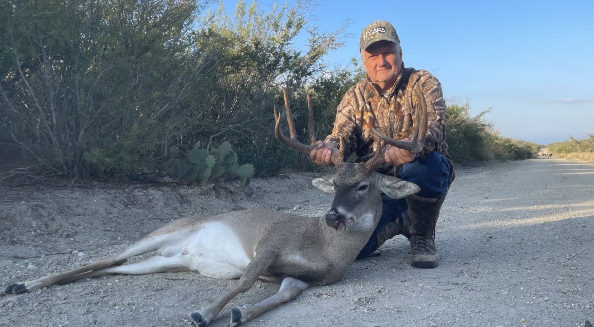 Man wearing camouflage jacket and cap kneeling on a dirt road, holding the antlers of a large buck deer lying on the ground.