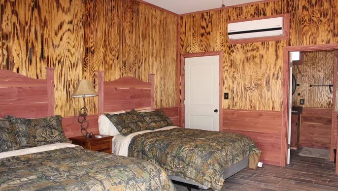 Wood-paneled bedroom with two beds, camouflage-patterned bedding, a nightstand with a lamp, a white door, and an air conditioner mounted on the wall.