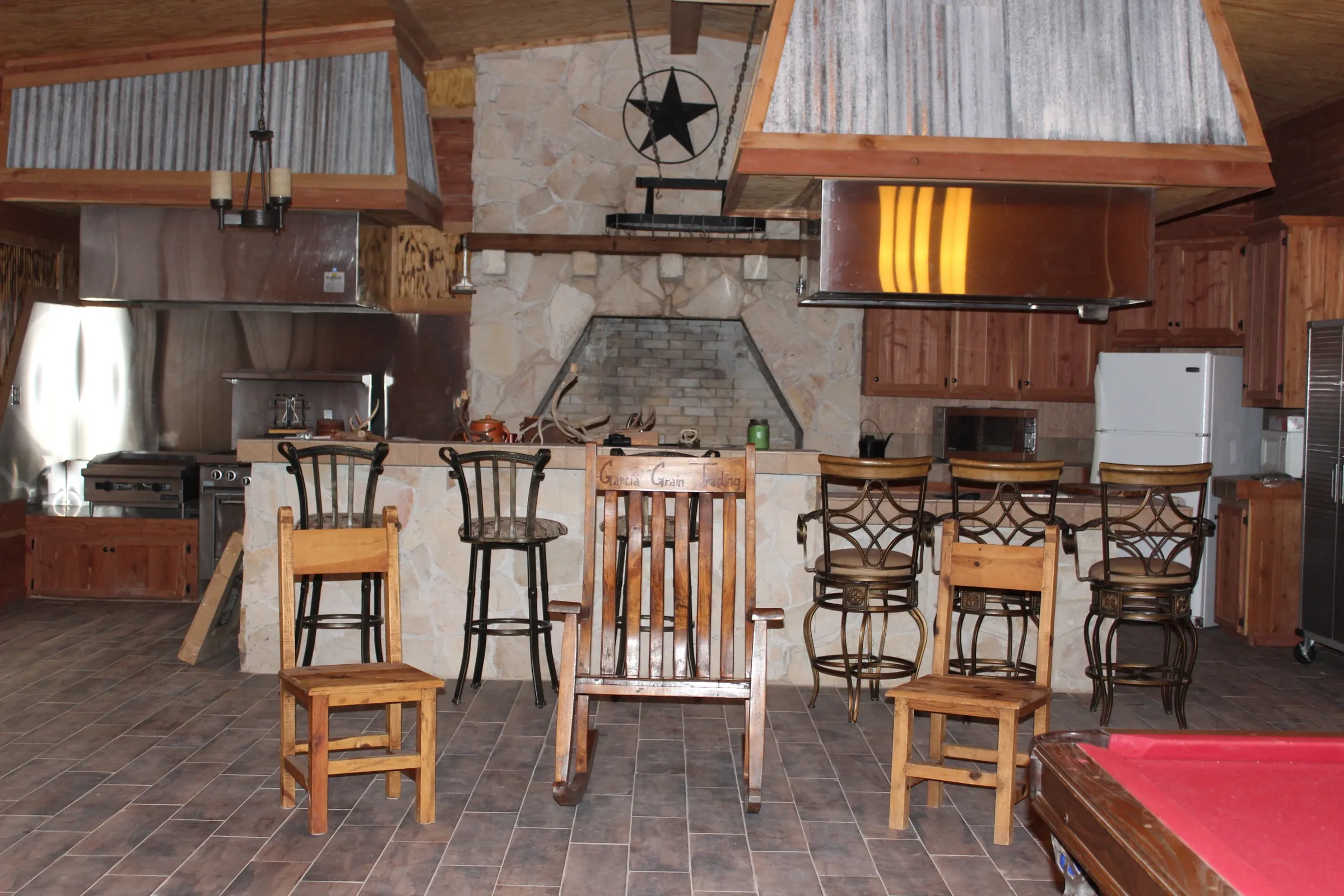 Rustic lodge kitchen with stone fireplace, wooden cabinetry, metal bar stools, wooden chairs, and a partial view of a red pool table.